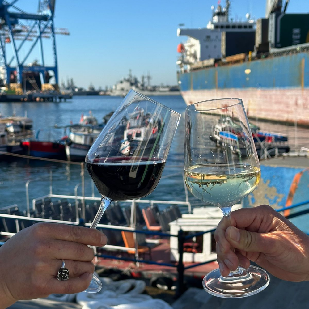 Dos copas de vino, una de tinto y otra de blanco, brindando frente al puerto de Valparaíso, con barcos, grúas y contenedores de fondo.