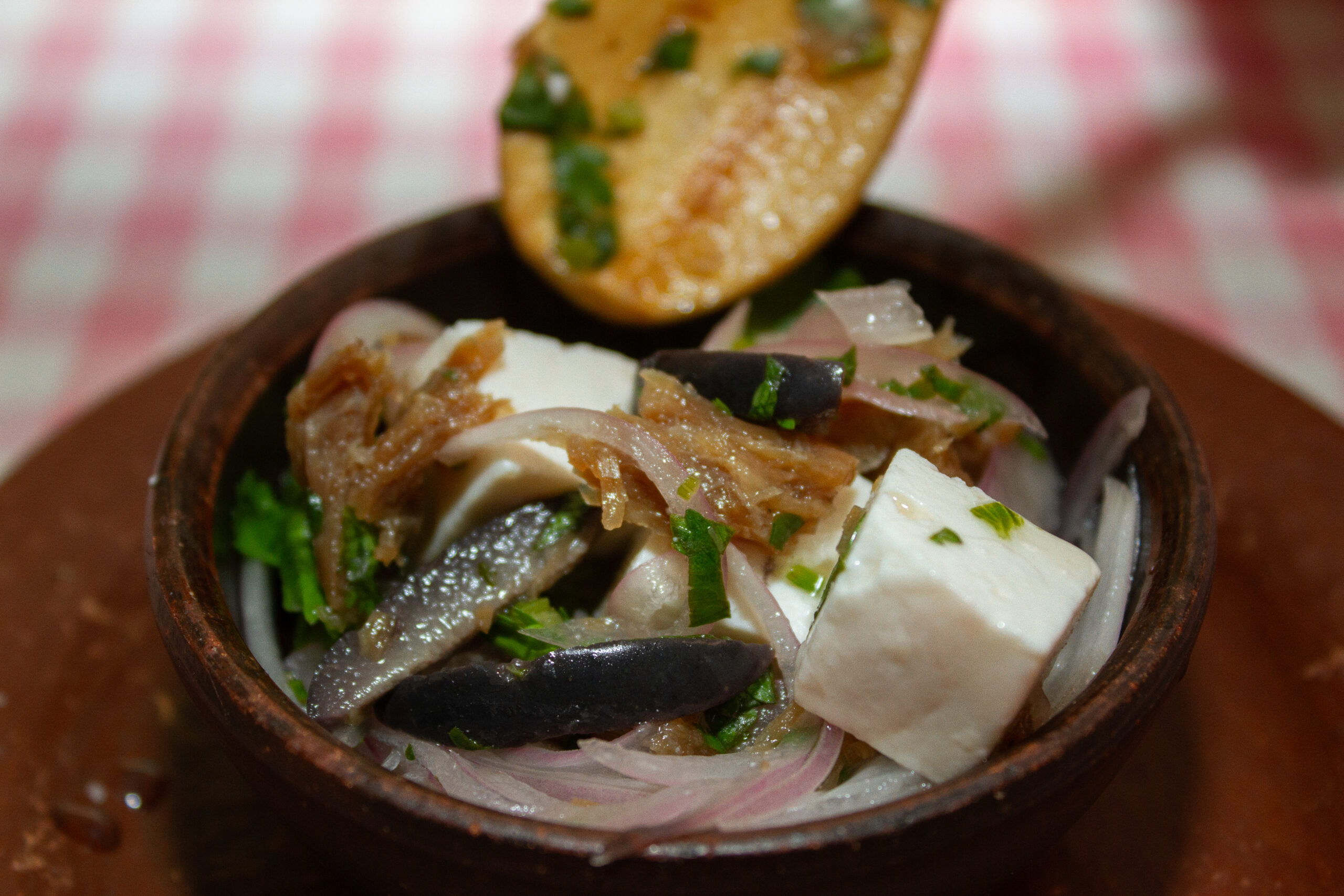 Mano sosteniendo una cuchara mientras se vierte una salsa cremosa de matico desde una jarra blanca sobre un plato de machas frescas, decoradas con cilantro y finas hierbas. La imagen captura el momento preciso en que la salsa cae suavemente, resaltando la textura y el color del plato en una presentación elegante y tradicional.